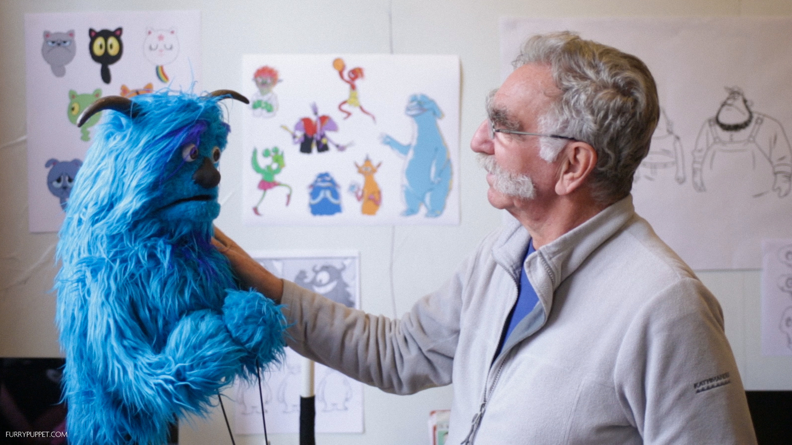 Furry Puppet Studio fabricator holding a blue furry marionette beside character design sketches